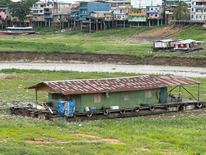 A simple home constructed of wood and metal sits alongside the edge of a river with a backdrop of the housing conditions of the city's outskirts in Manaus, the capital city of the Brazilian state of Amazonas.