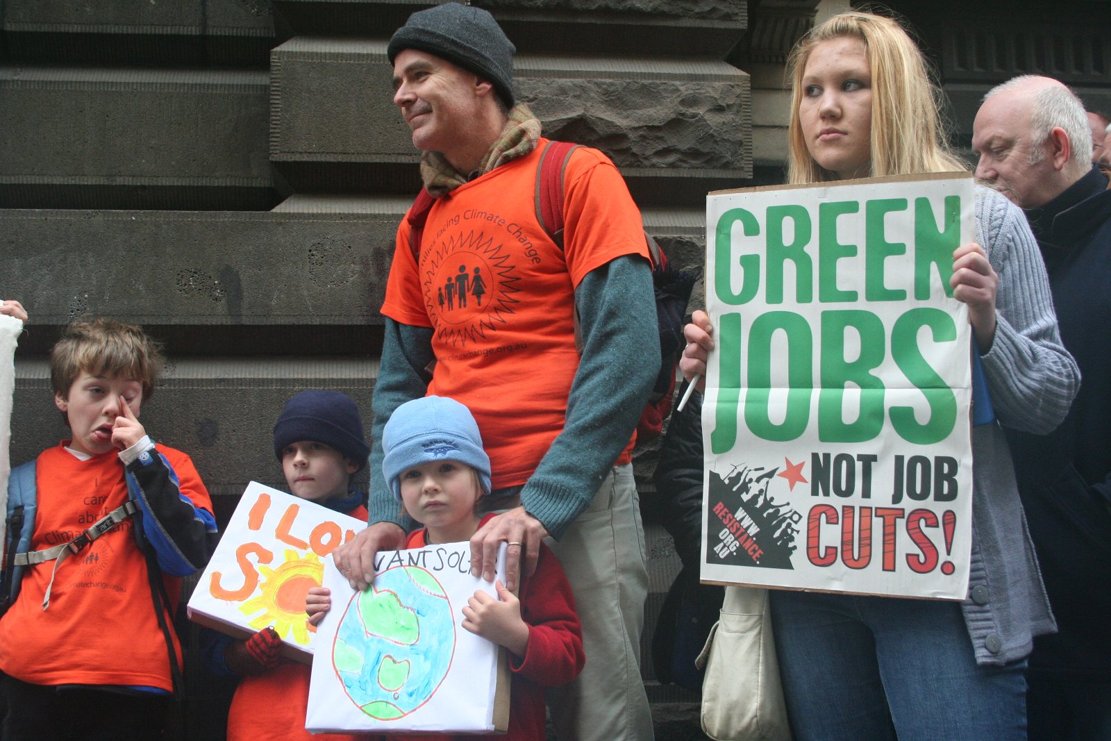 Woman holds up a sign that says “Green Jobs Not Job Cuts!”  at a rally for climate change in Melbourne with a man and three young children. 