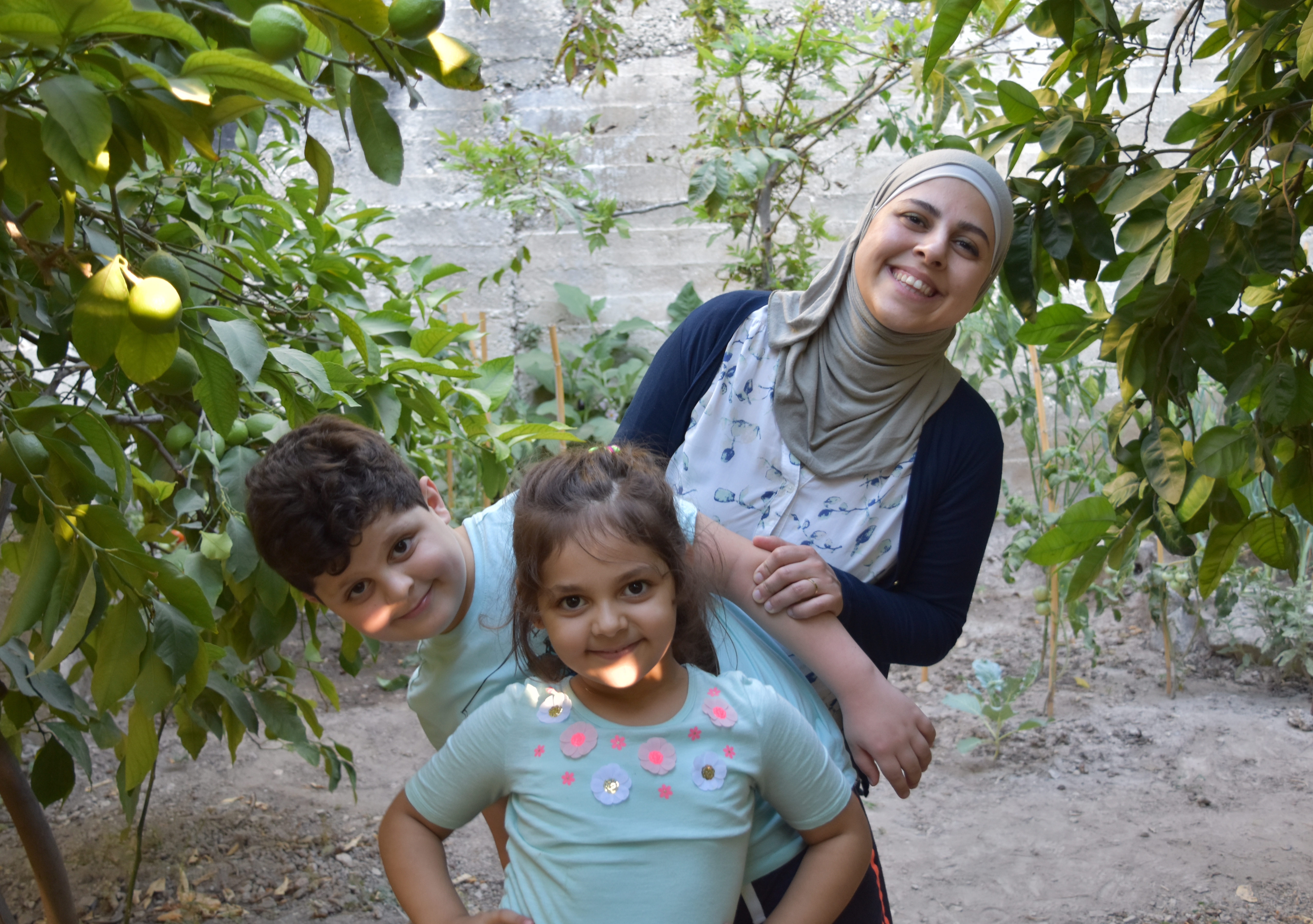 Photo of a woman and two young children smiling on a farm in front of trees. 