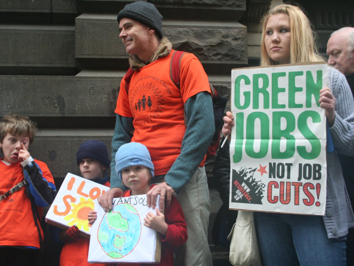Woman holds up a sign that says “Green Jobs Not Job Cuts!” at a rally with a man and three children 