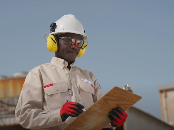 A man wearing a hardhat and noise protection ear muffs. He his holding a clipboard outdoors.