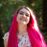 Photo of a white girl with long bright pink hair, smiling big at the camera with trees and greenery behind them, wearing a long floral dress.