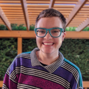Photo of a white person with short brown hair smiling at the camera; they are wearing a striped sweater and blue glasses, and have a nose ring.